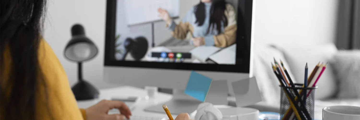 Pessoa assiste aula online no computador enquanto anota em caderno em ambiente de estudo organizado.