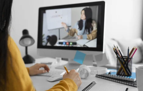Pessoa assiste aula online no computador enquanto anota em caderno em ambiente de estudo organizado.