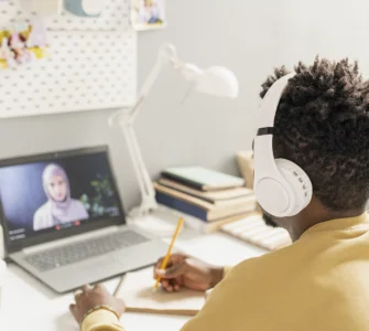 Pessoa com fones de ouvido estuda online, assistindo aula em laptop e fazendo anotações em caderno.