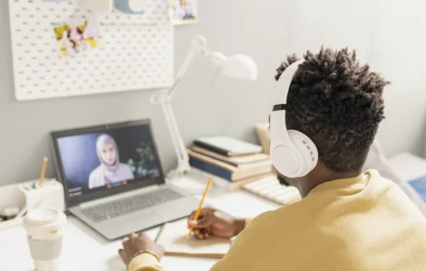 Pessoa com fones de ouvido estuda online, assistindo aula em laptop e fazendo anotações em caderno.