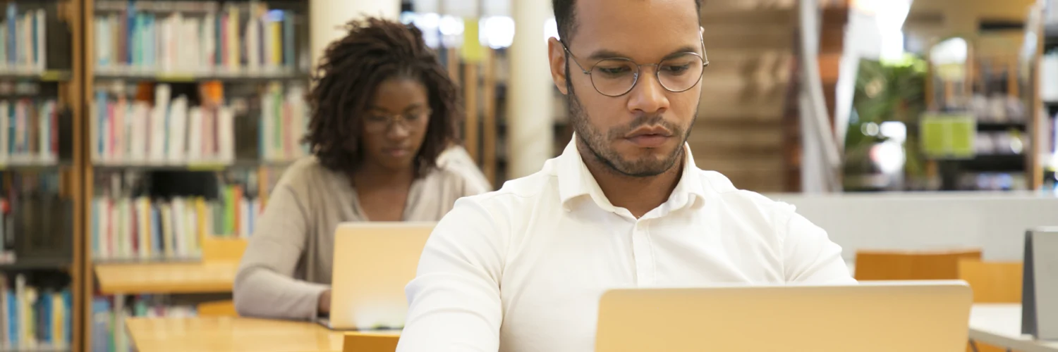 Homem negro com camisa branca e óculos trabalha em um laptop, com uma mulher ao fundo em uma biblioteca.