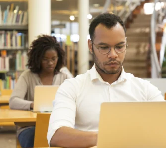Homem negro com camisa branca e óculos trabalha em um laptop, com uma mulher ao fundo em uma biblioteca.