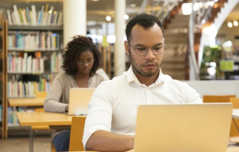 Homem negro com camisa branca e óculos trabalha em um laptop, com uma mulher ao fundo em uma biblioteca.