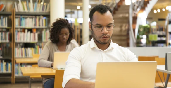 Homem negro com camisa branca e óculos trabalha em um laptop, com uma mulher ao fundo em uma biblioteca.