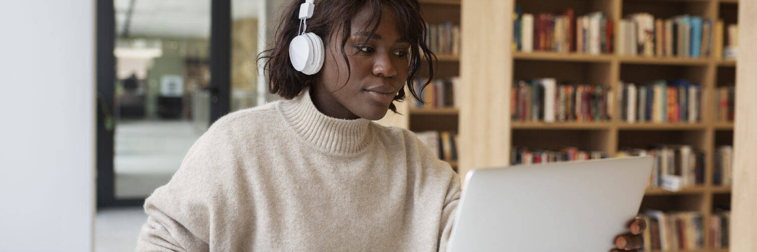 Mulher de pele negra com fones de ouvido brancos, usando um laptop em uma mesa com cadernos, em uma biblioteca.