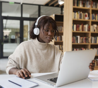 Mulher de pele negra com fones de ouvido brancos, usando um laptop em uma mesa com cadernos, em uma biblioteca.