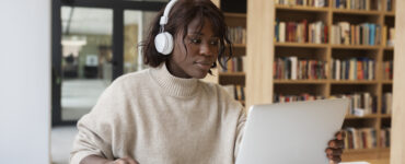 Mulher de pele negra com fones de ouvido brancos, usando um laptop em uma mesa com cadernos, em uma biblioteca.