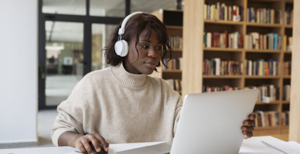 Mulher de pele negra com fones de ouvido brancos, usando um laptop em uma mesa com cadernos, em uma biblioteca.