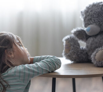 Menina observa pensativa seu ursinho de pelúcia sobre a mesa, em ambiente suave e iluminado.