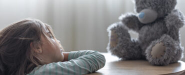 Menina observa pensativa seu ursinho de pelúcia sobre a mesa, em ambiente suave e iluminado.