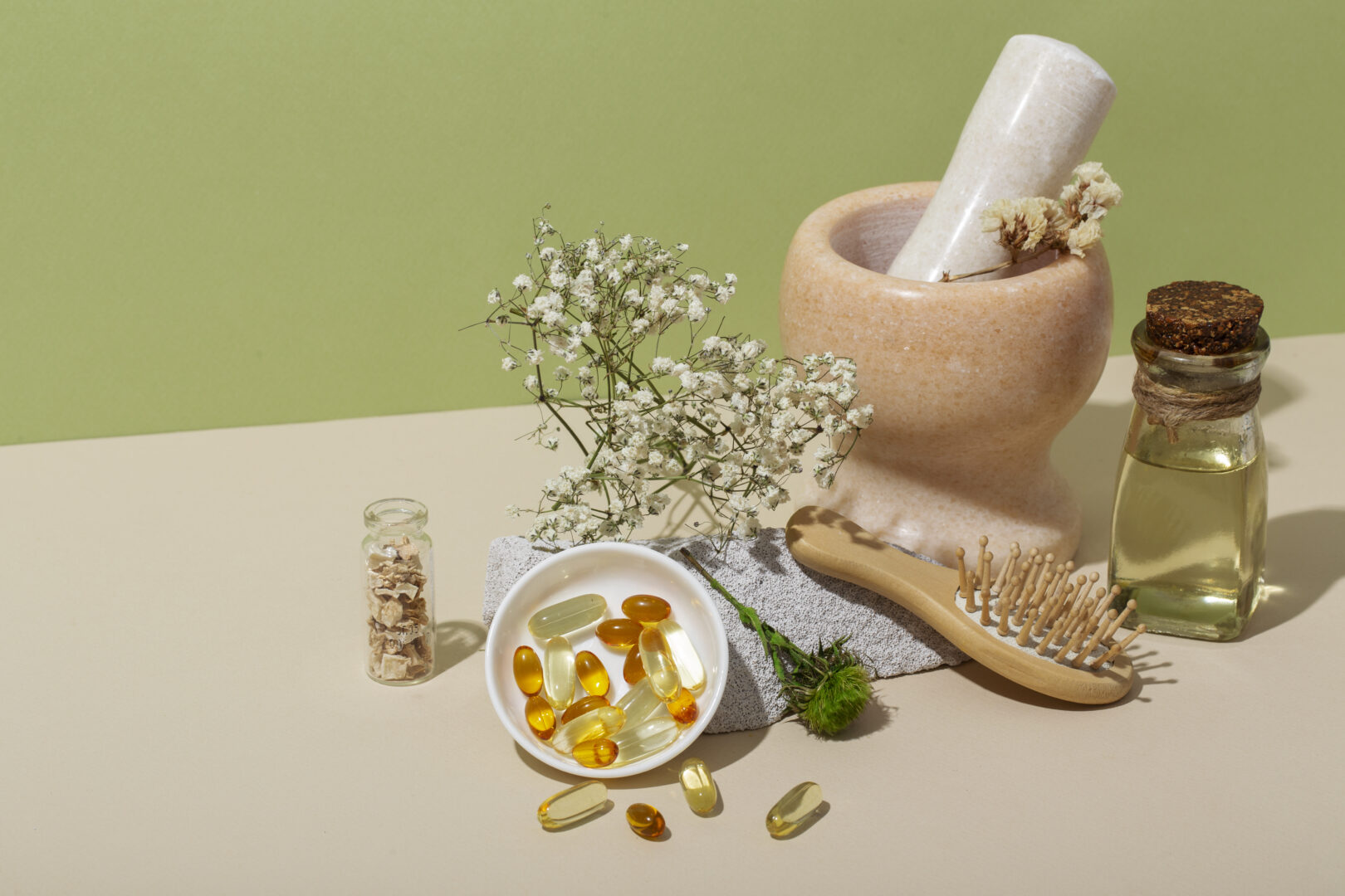Cápsulas, frascos com óleo, flores secas e pilão de mármore sobre fundo verde e bege.