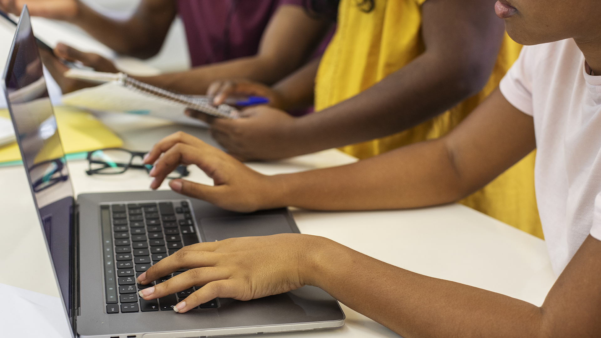 Mãos de estudantes digitando em um laptop e escrevendo em caderno durante atividade em grupo.