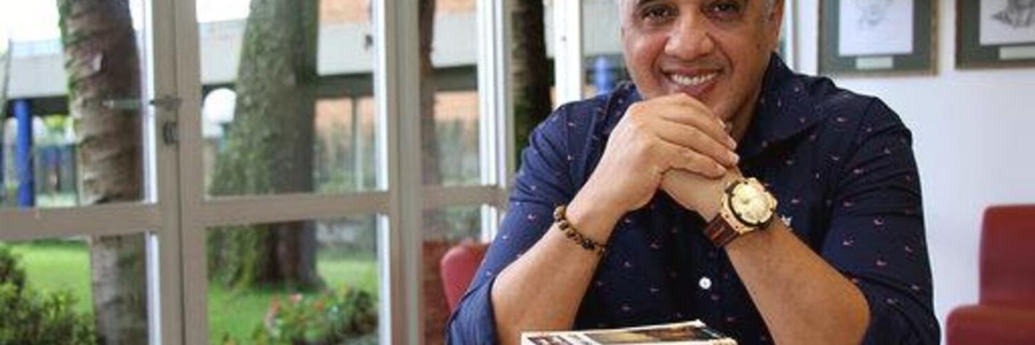 Imagem do entrevistado, João Jonas, sorrindo para a câmera com uma pilha de livros em sua frente.