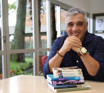 Imagem do entrevistado, João Jonas, sorrindo para a câmera com uma pilha de livros em sua frente.