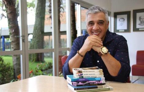 Imagem do entrevistado, João Jonas, sorrindo para a câmera com uma pilha de livros em sua frente.