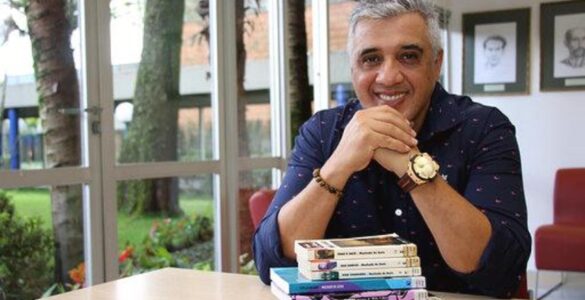 Imagem do entrevistado, João Jonas, sorrindo para a câmera com uma pilha de livros em sua frente.