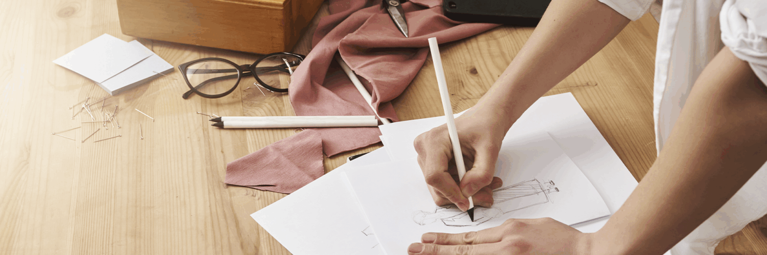 Mãos desenhando croqui de moda. Mesa com máquina de costura vintage, tesoura, tecido rosa, óculos e alfinetes.