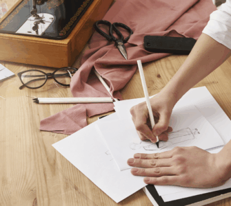 Mãos desenhando croqui de moda. Mesa com máquina de costura vintage, tesoura, tecido rosa, óculos e alfinetes.