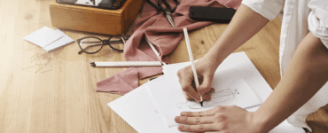 Mãos desenhando croqui de moda. Mesa com máquina de costura vintage, tesoura, tecido rosa, óculos e alfinetes.