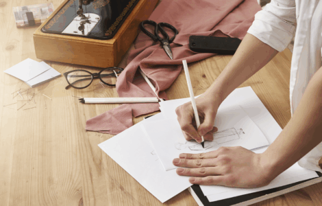Mãos desenhando croqui de moda. Mesa com máquina de costura vintage, tesoura, tecido rosa, óculos e alfinetes.