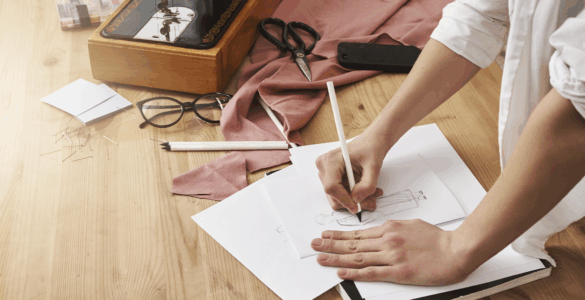 Mãos desenhando croqui de moda. Mesa com máquina de costura vintage, tesoura, tecido rosa, óculos e alfinetes.