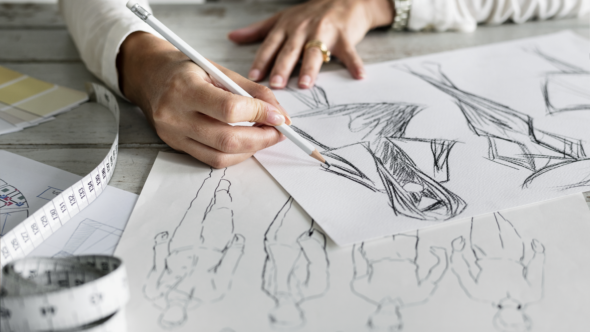Artista desenhando croquis de moda com lápis e régua, cercado de tecidos e esquemas de roupas, em um ambiente de ateliê.